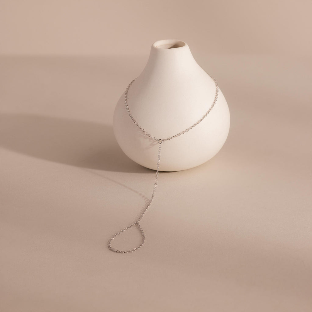 Delicate silver hand chain bracelet elegantly draped over a small matte white ceramic vase, casting a soft shadow.