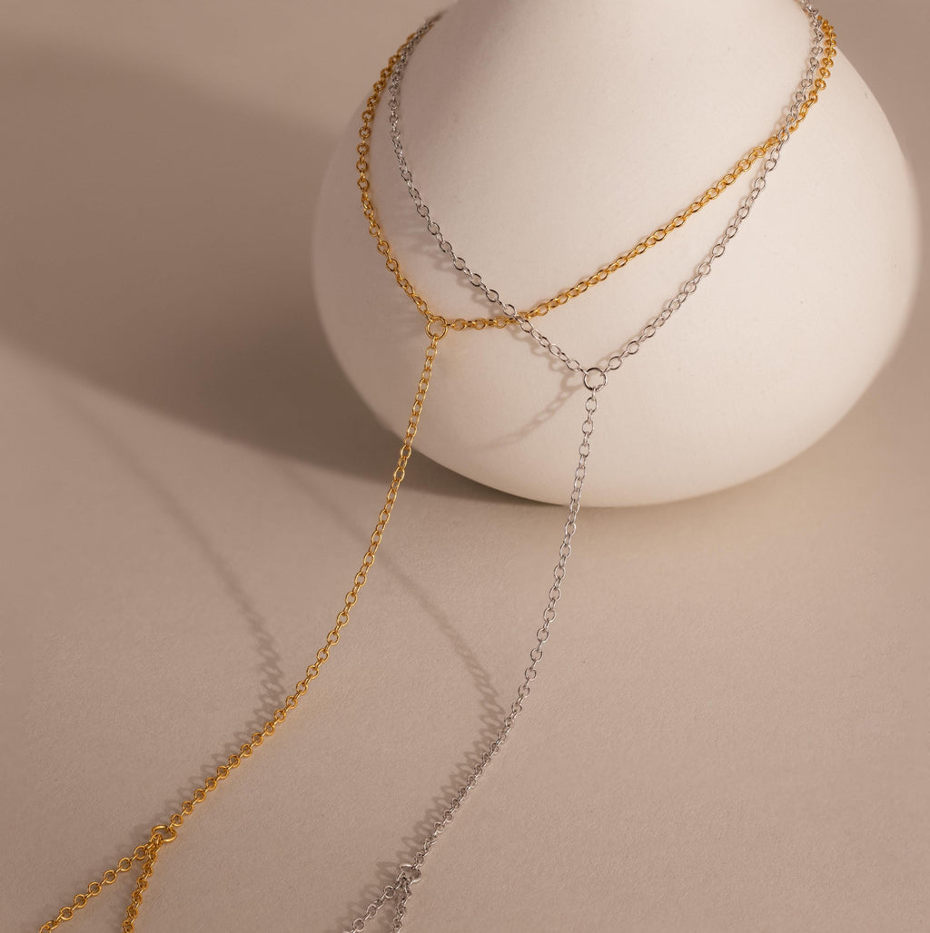 Two-tone dainty hand chain bracelets in gold and silver draped over a minimal ceramic vase, with delicate chain strands creating elegant shadows on a neutral surface.