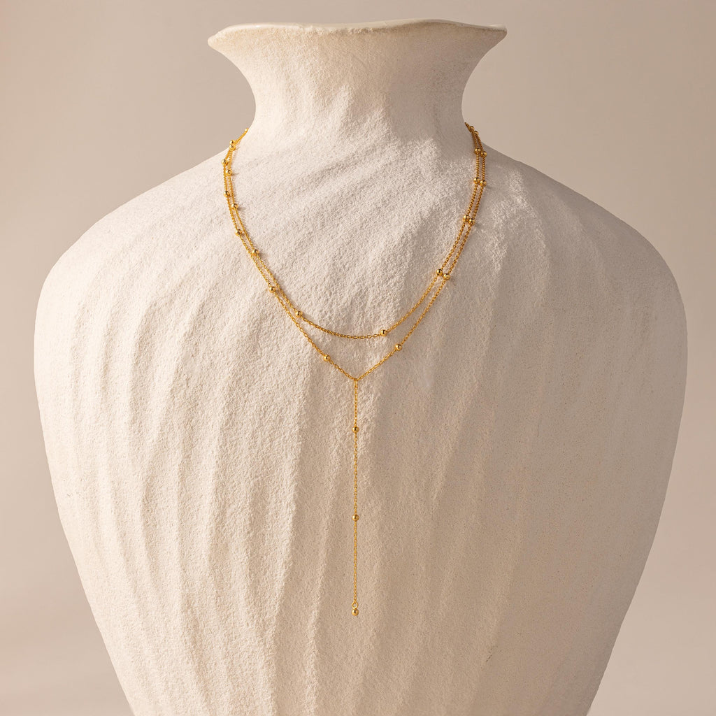 Gold satellite lariat necklace displayed on a sculptural ivory vase, showing full length and layered design.