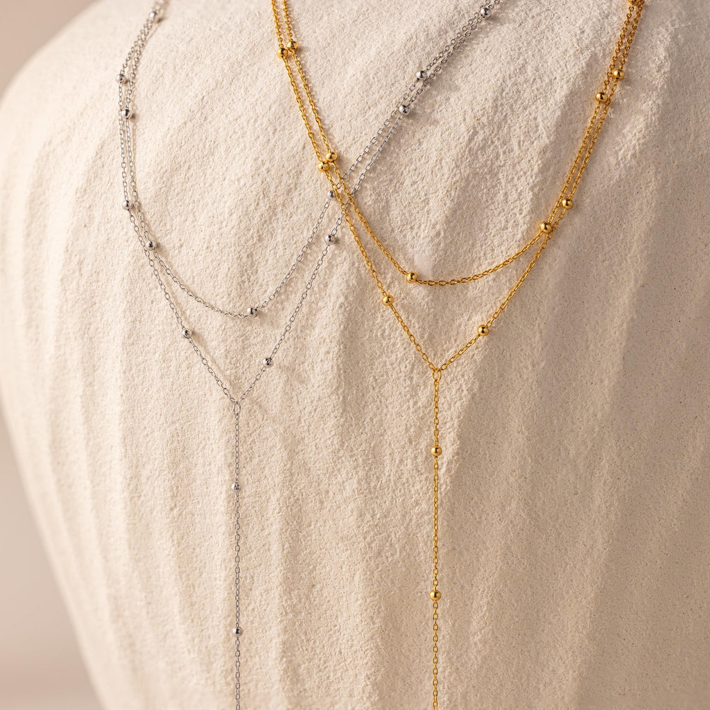 Side-by-side display of two Duo Satellite Lariat Necklaces—one in silver and one in gold—draped elegantly over a textured off-white vase.