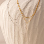 Side-by-side display of two Duo Satellite Lariat Necklaces—one in silver and one in gold—draped elegantly over a textured off-white vase.