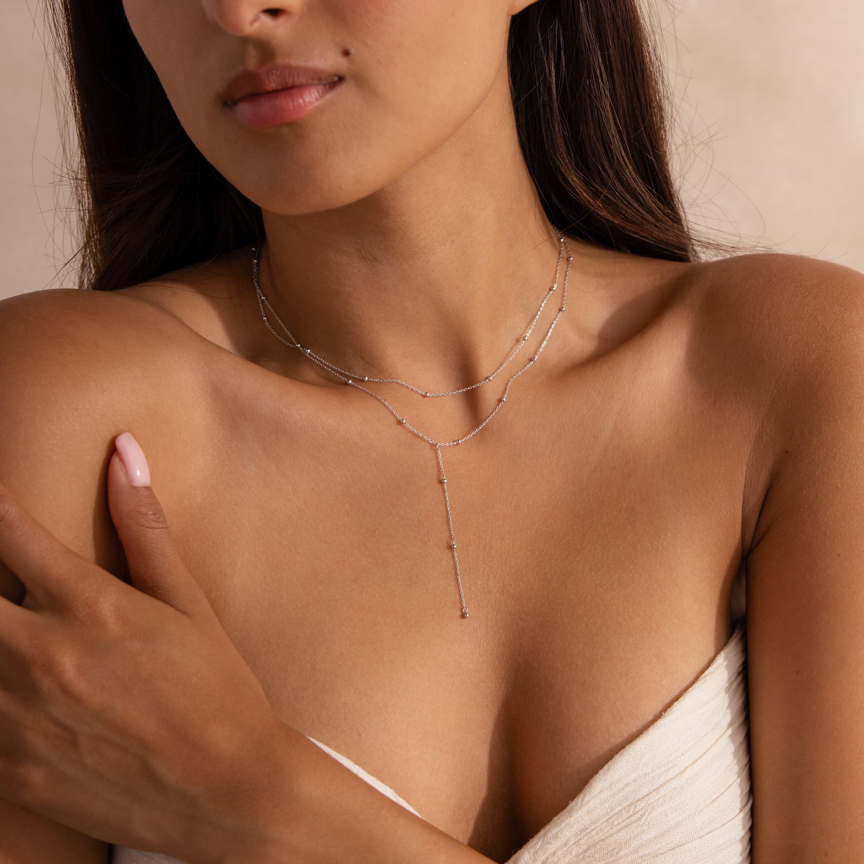 A woman wearing a silver layered lariat necklace with delicate satellite chain details, styled with an off-white strapless top.