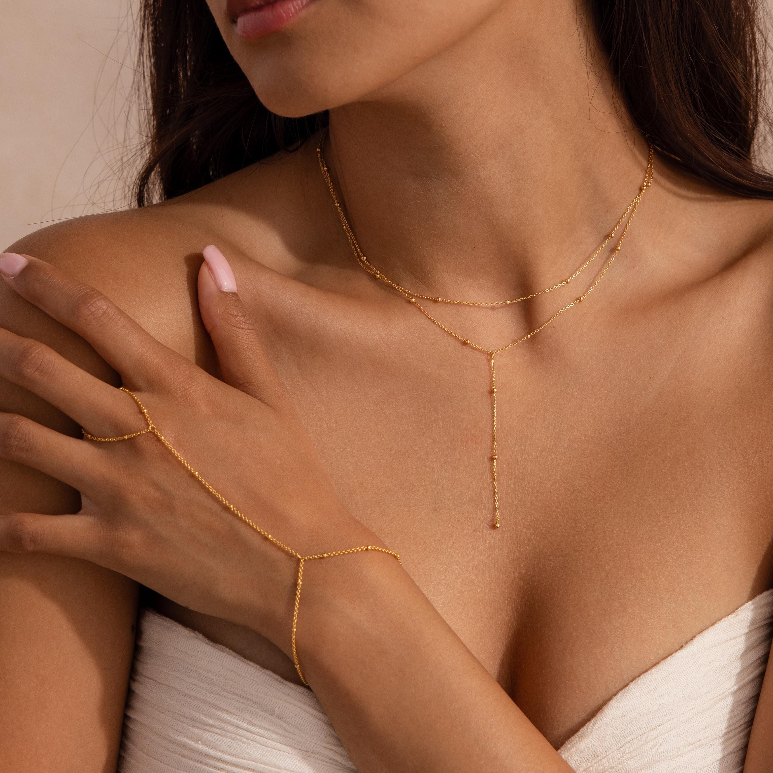 Woman wearing a layered gold lariat necklace and matching gold hand chain, styled with a strapless white top.