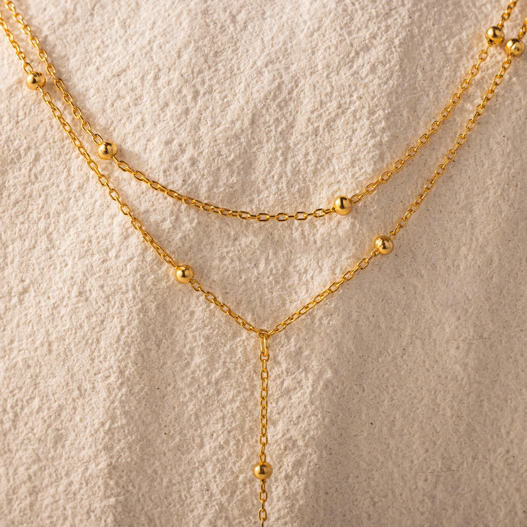 Close-up of the necklace’s gold satellite bead chain against a textured cream background.