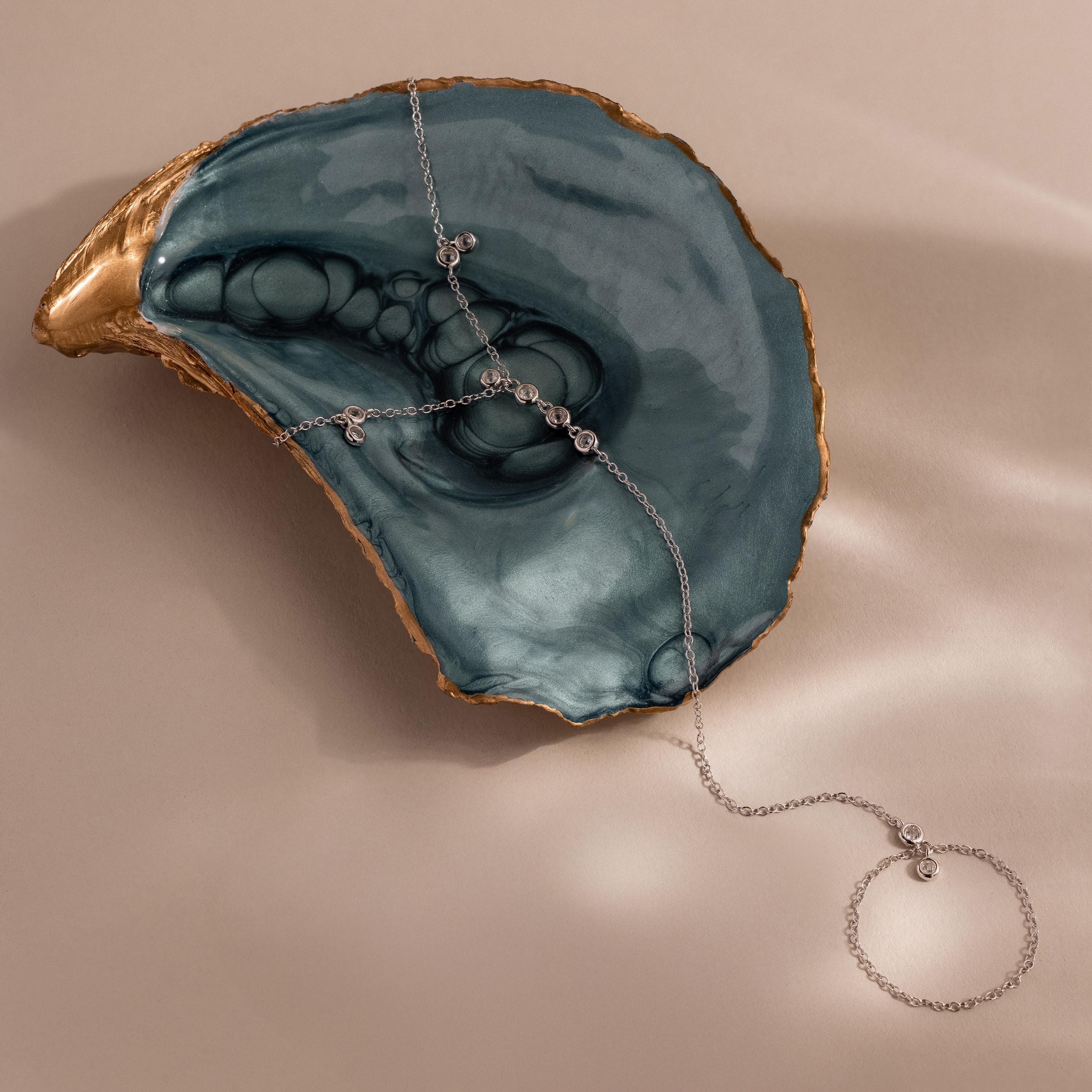 Close-up of a silver hand chain bracelet artfully draped over a teal marbled oyster shell with gold trim.