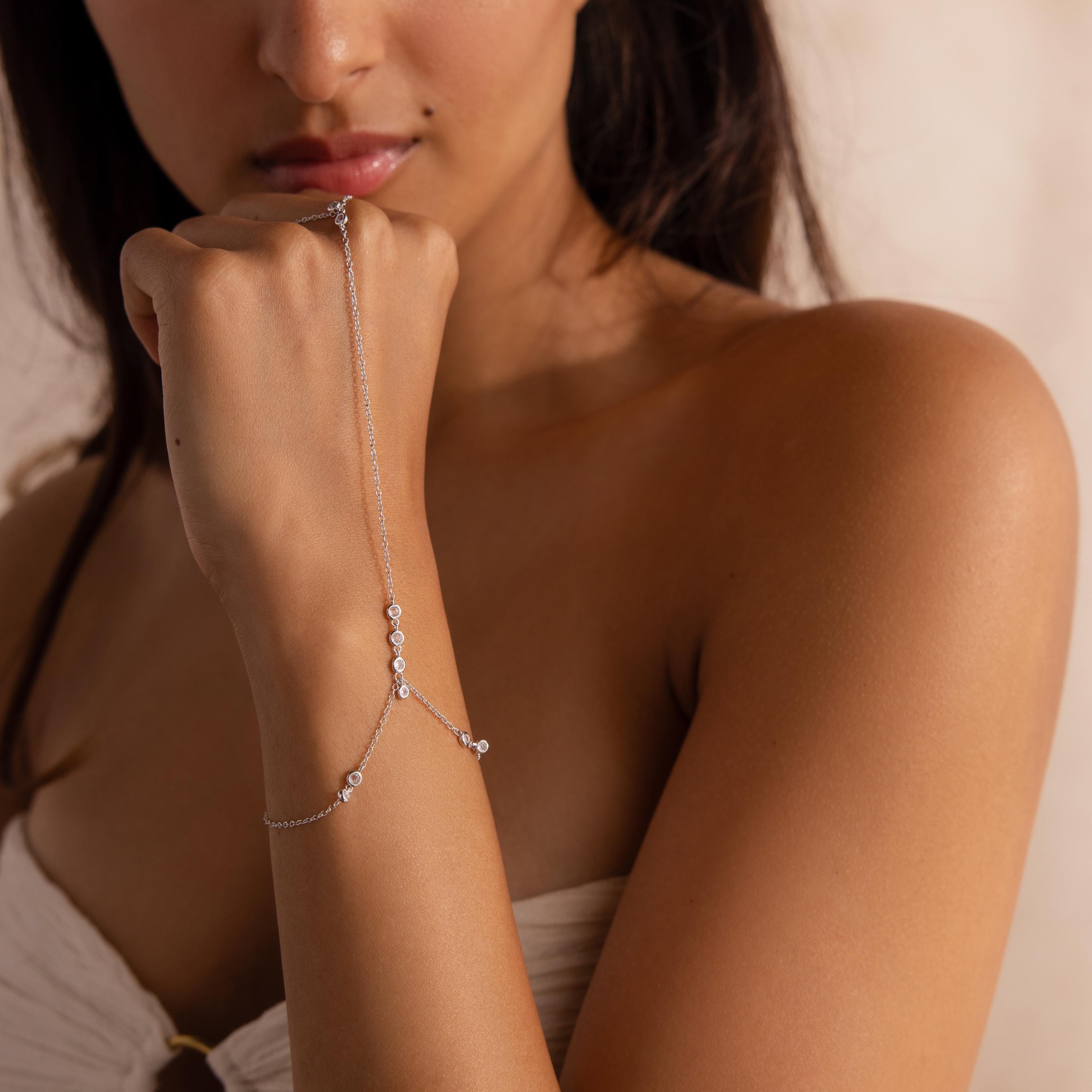 Woman wearing a dainty silver hand chain bracelet that runs from the wrist to a central ring on her finger, styled with a strapless cream top.