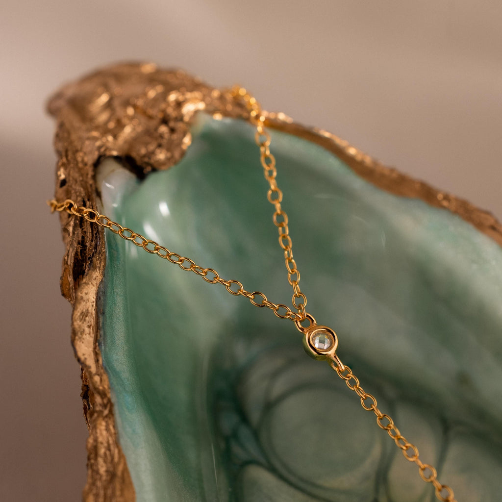 Close-up of a gold hand chain draped over a green and gold seashell dish, showcasing a single bezel-set diamond.