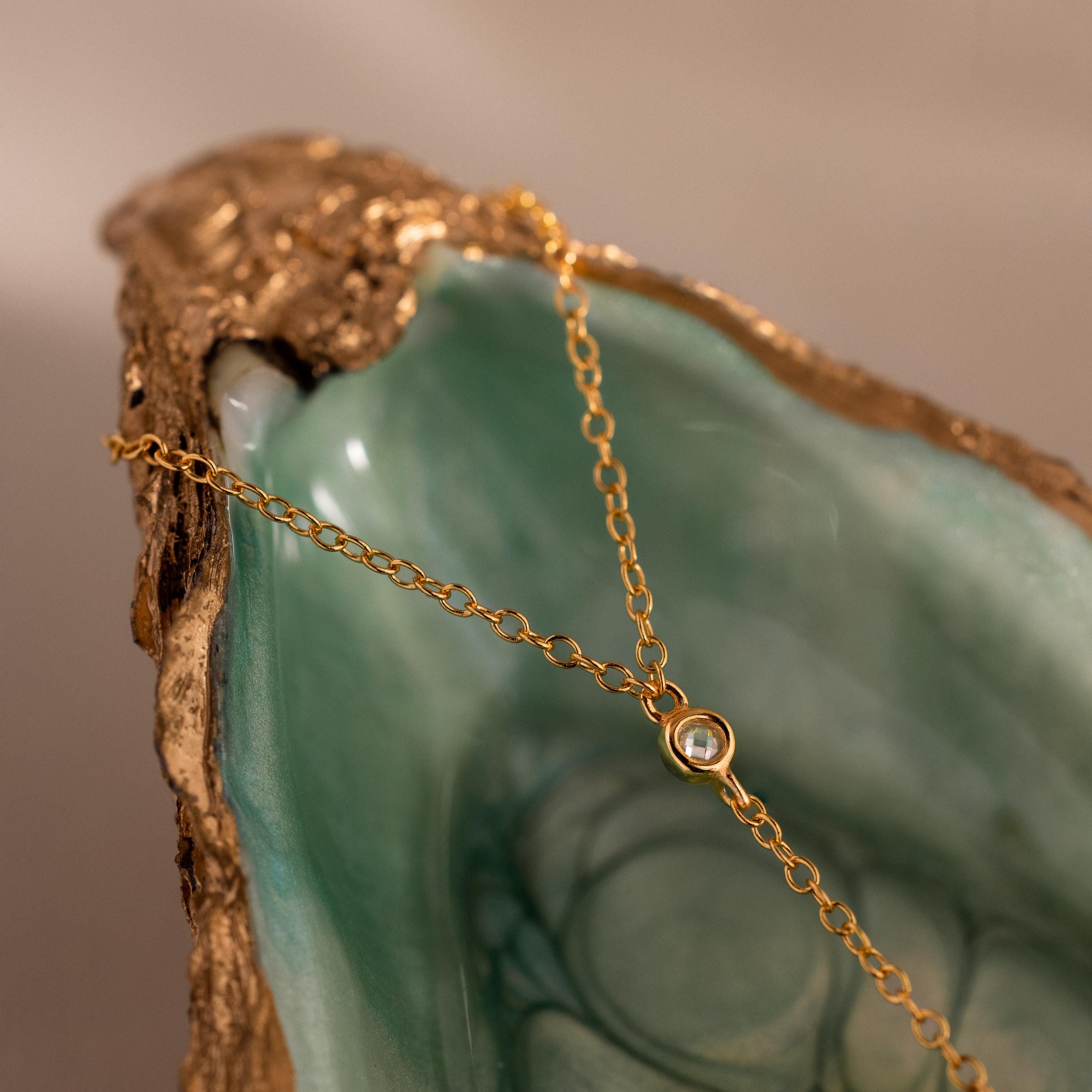 Close-up of a gold hand chain draped over a green and gold seashell dish, showcasing a single bezel-set diamond.