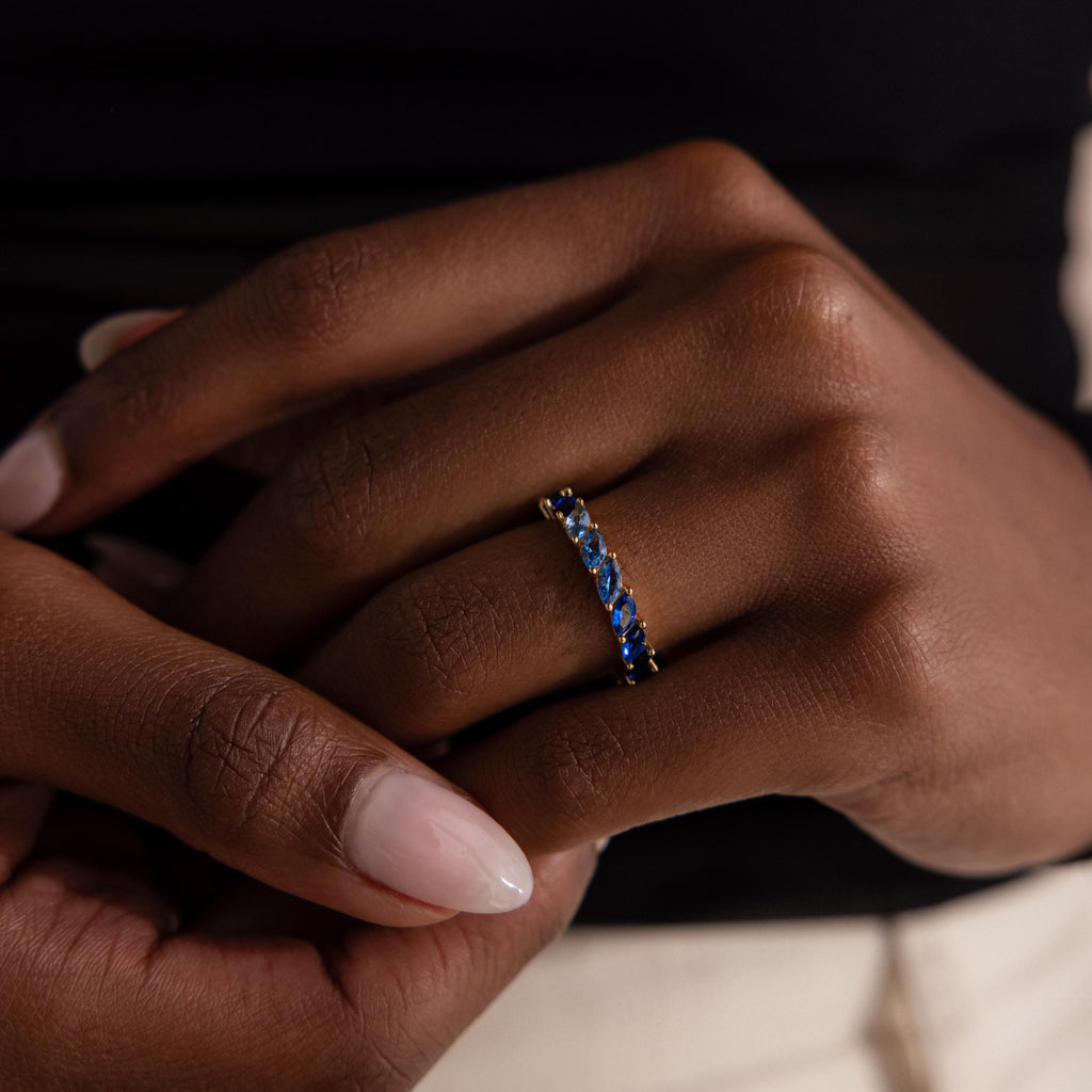 Close up of a hand wearing a gold eternity band with marquise cut stones in an ombre gradient from deep sapphire to icy blue, catching light with a delicate shimmer.