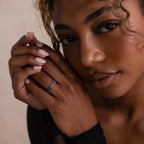 Model wears a gold marquise cut eternity ring on her index finger, showcasing a gradient of blue stones that shift from deep navy to icy pale tones. Hands are gently posed near her face in soft lighting.