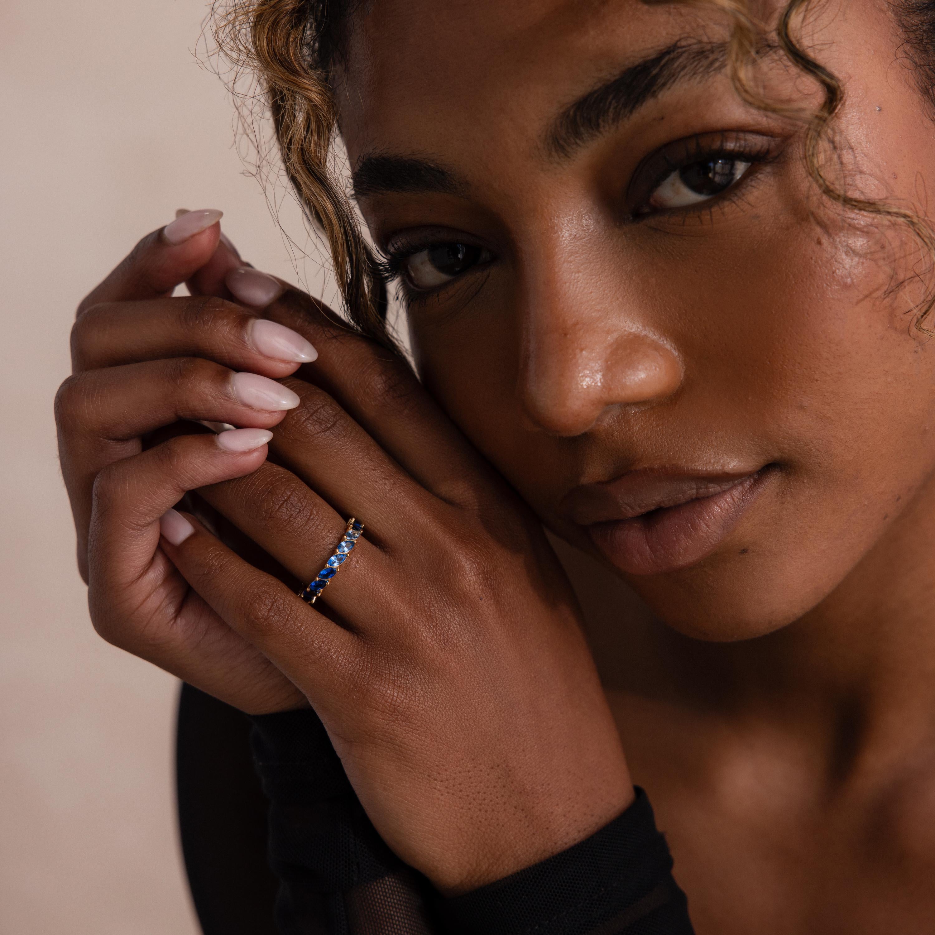 Model wears a gold marquise cut eternity ring on her index finger, showcasing a gradient of blue stones that shift from deep navy to icy pale tones. Hands are gently posed near her face in soft lighting.