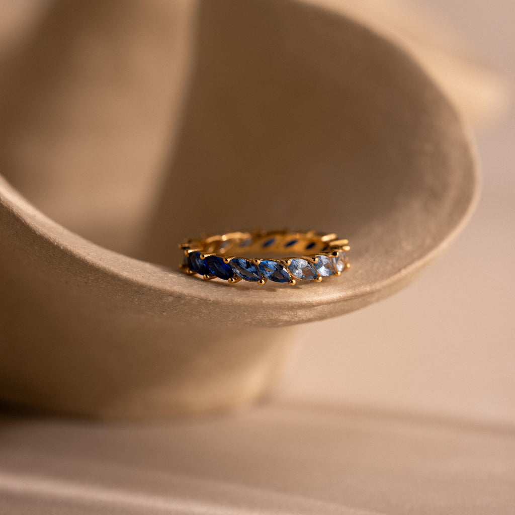 Gold eternity band with marquise cut stones in a gradient of deep to light blue rests inside a sculptural ceramic dish, softly lit to highlight the ombre effect and prong setting.