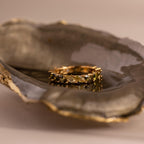 Gold ring with marquise-cut green ombré stones rests inside a dark, glossy oyster shell with metallic gold edging. The gems fade from deep olive to light yellow-green.