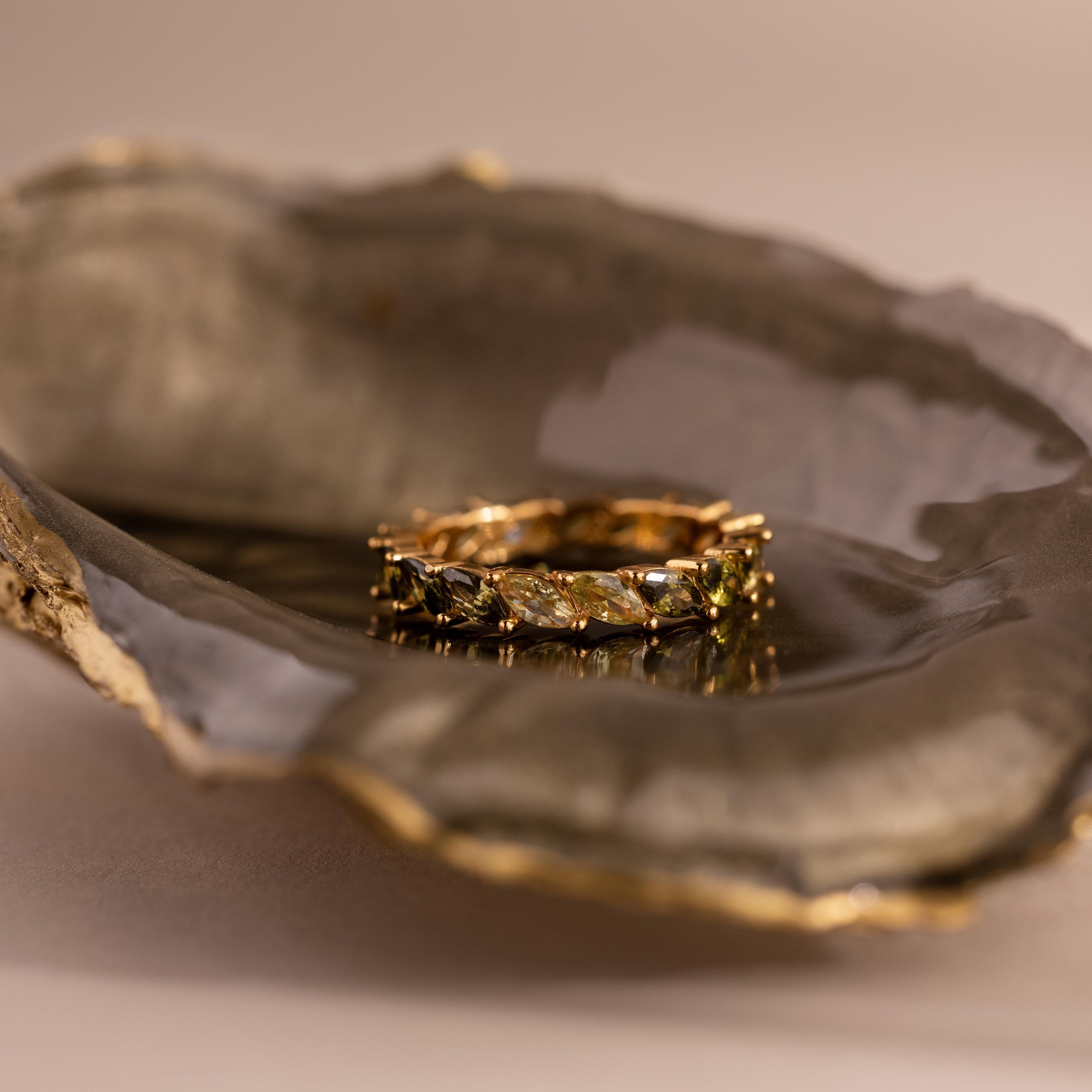 Gold ring with marquise-cut green ombré stones rests inside a dark, glossy oyster shell with metallic gold edging. The gems fade from deep olive to light yellow-green.