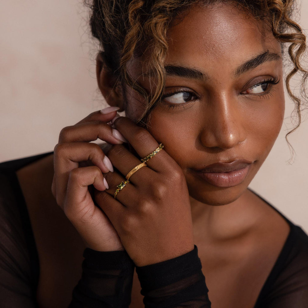 Model poses with hands gently resting on her face, wearing three gold rings including a marquise cut green ombré eternity band. The soft lighting and sheer black sleeves highlight the ring’s elegant sparkle.