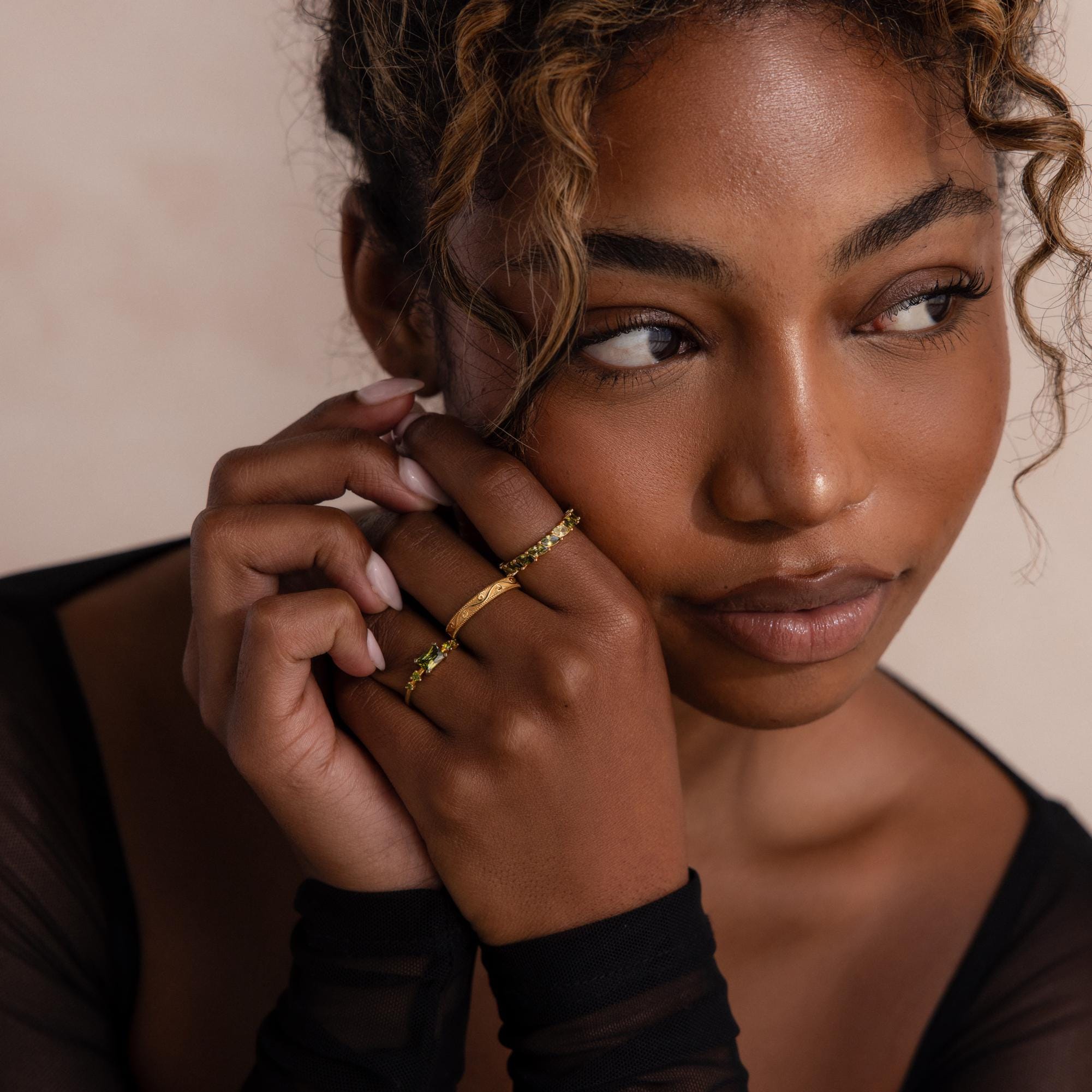 Model poses with hands gently resting on her face, wearing three gold rings including a marquise cut green ombré eternity band. The soft lighting and sheer black sleeves highlight the ring’s elegant sparkle.
