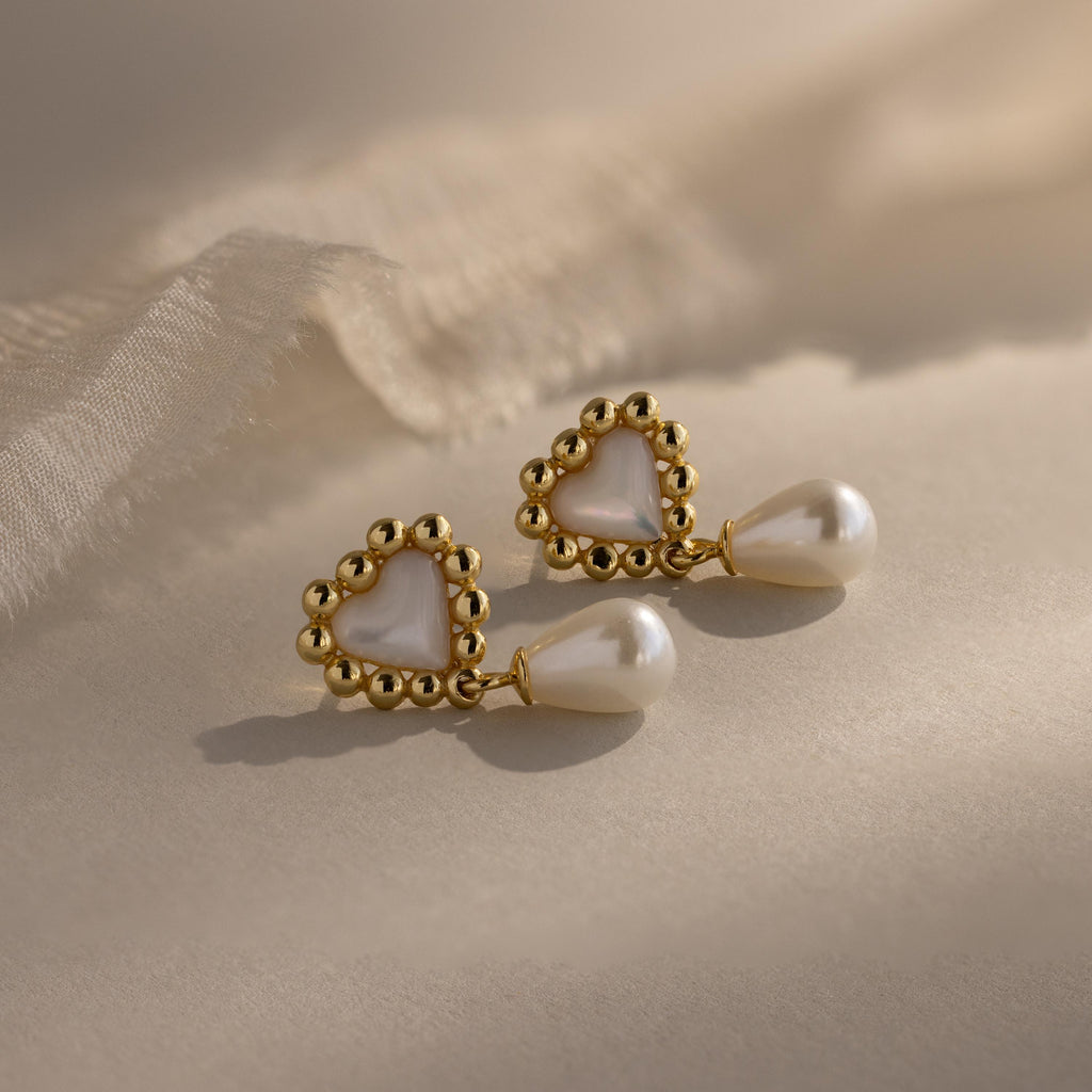 Flat lay of gold beaded heart earrings with pearl drops, arranged in warm sunlight on a beige fabric surface.