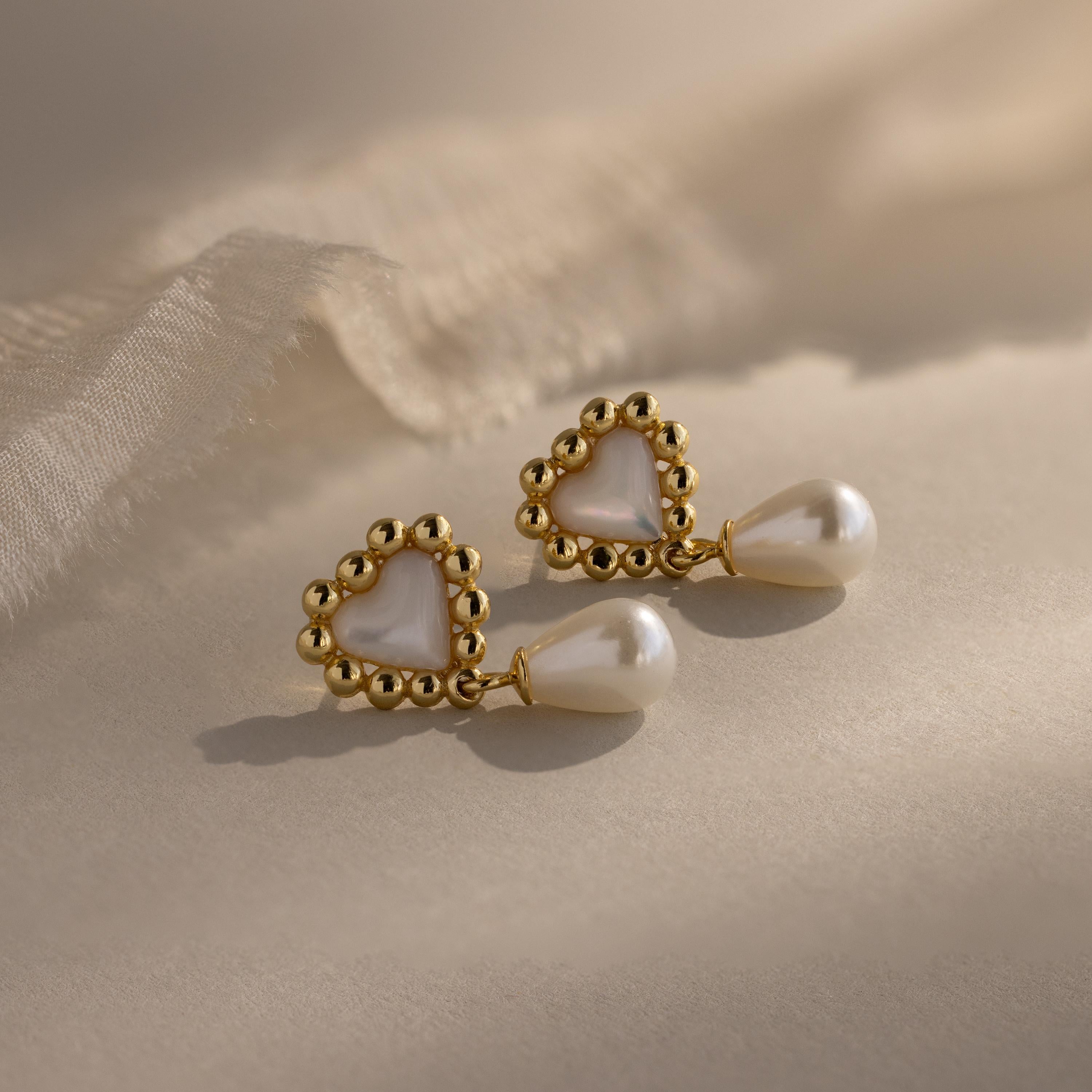 Flat lay of gold beaded heart earrings with pearl drops, arranged in warm sunlight on a beige fabric surface.