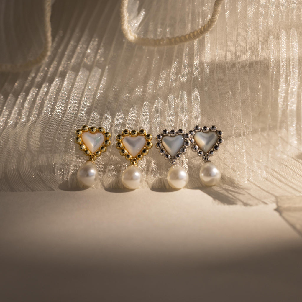 Four pairs of beaded pearl heart earrings—two in gold, two in silver—arranged in a row on pleated fabric with soft shadows.