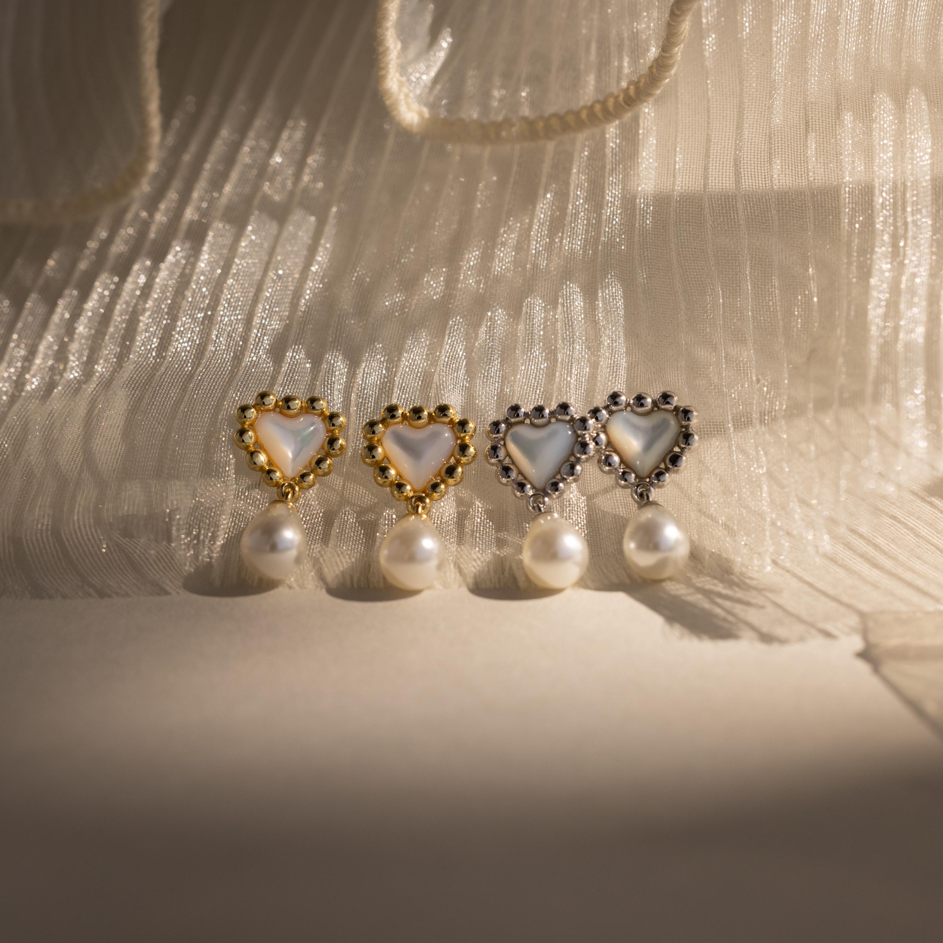 Four pairs of beaded pearl heart earrings—two in gold, two in silver—arranged in a row on pleated fabric with soft shadows.