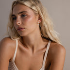 Model seated in a relaxed pose wearing gold pearl heart earrings, styled with a neutral-toned tank top.