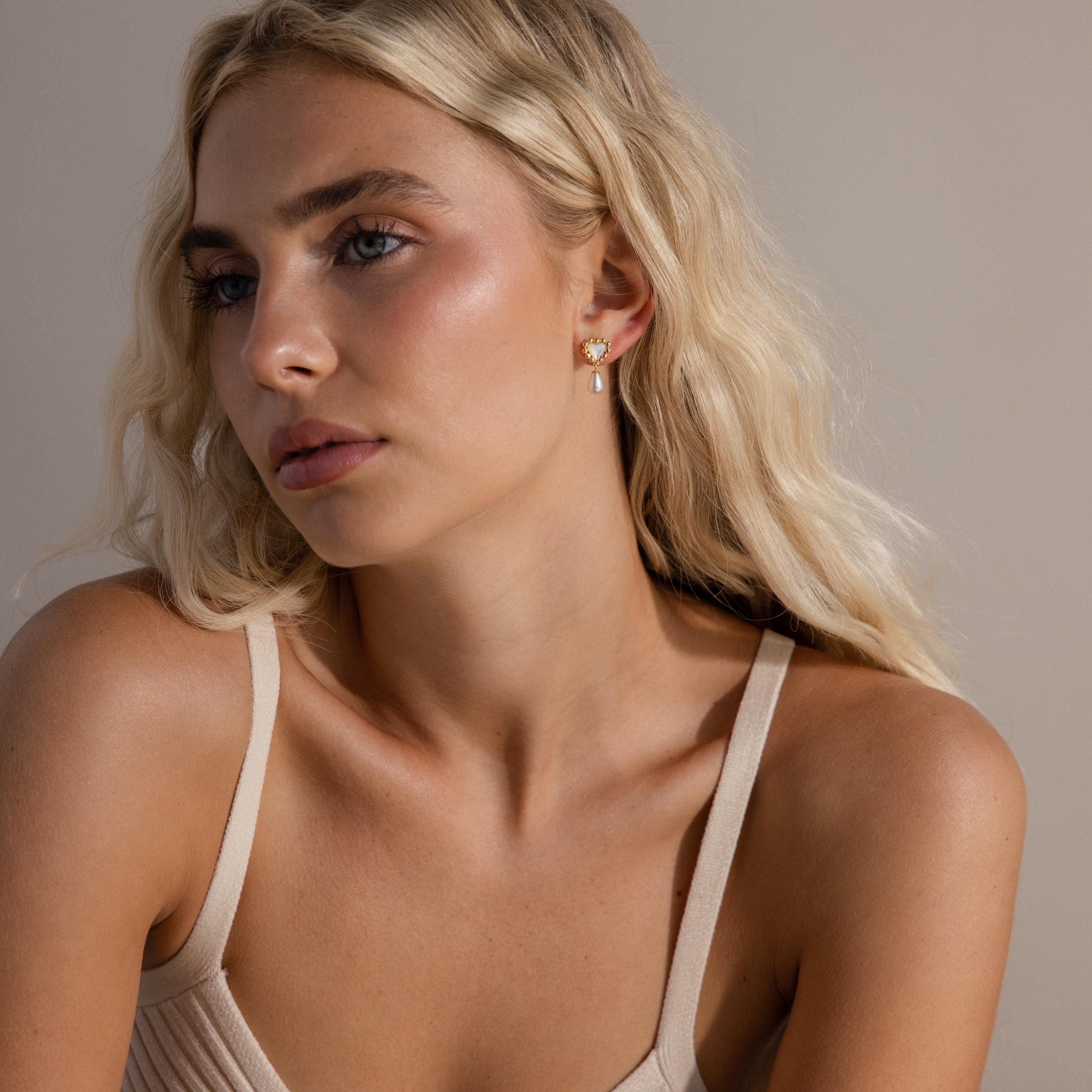 Model seated in a relaxed pose wearing gold pearl heart earrings, styled with a neutral-toned tank top.