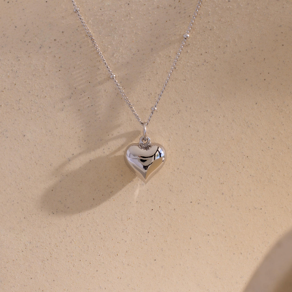 The white gold version of the Puff Heart Pendant Necklace displayed against a sand-colored background.