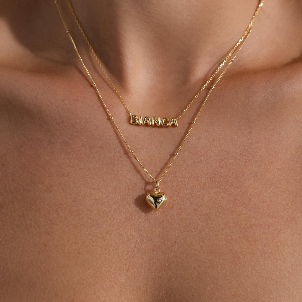 A layered necklace styling shot: the gold Puff Heart Pendant hangs beneath a second necklace that spells “BIANCA” in bold gold letters. Both necklaces sit perfectly centered on the model’s chest, creating a playful and personalized layered look.