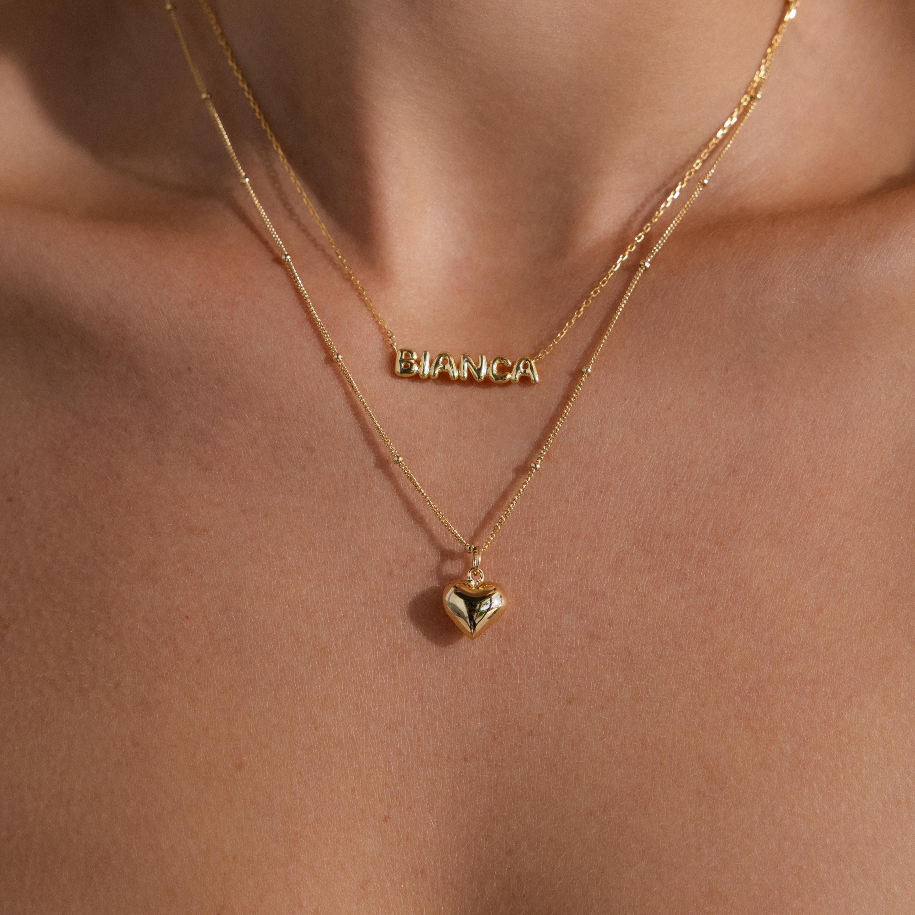 A layered necklace styling shot: the gold Puff Heart Pendant hangs beneath a second necklace that spells “BIANCA” in bold gold letters. Both necklaces sit perfectly centered on the model’s chest, creating a playful and personalized layered look.