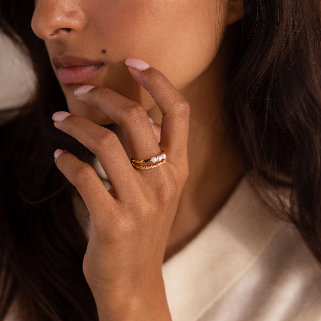 Woman wearing a duo band ring featuring a gold band with three small pearls and a pavé crystal band, styled with a soft cream top.