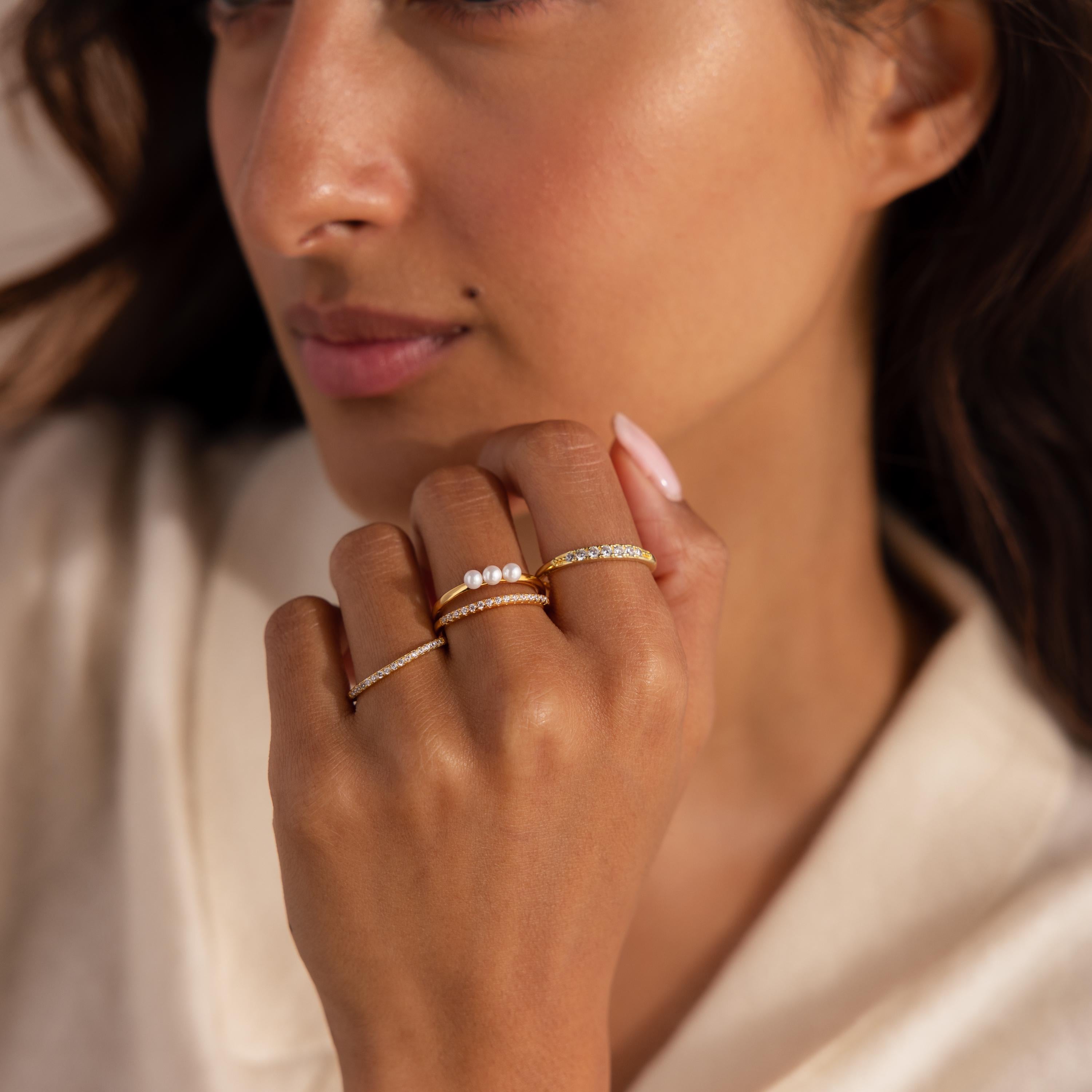 Close up of a woman wearing multiple gold rings on her fingers, including one with three small pearls and others with pavé set crystals, against a soft neutral background.