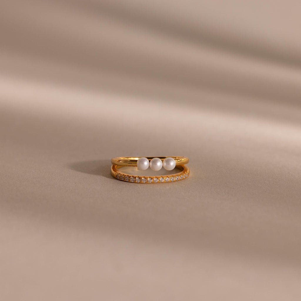 Gold duo ring featuring one band with three round white pearls and a second slim band with pavé crystals, displayed on a smooth beige surface.