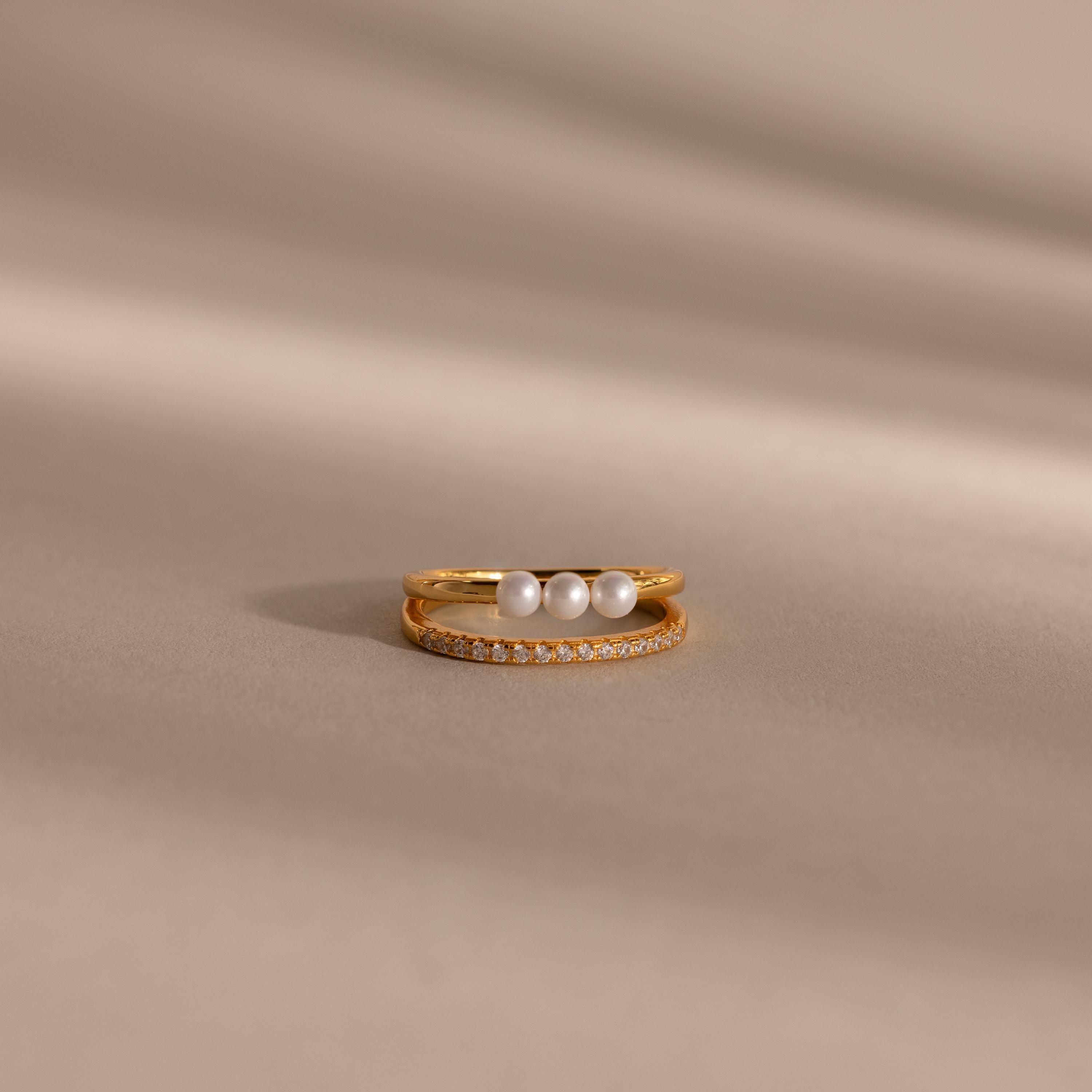 Gold duo ring featuring one band with three round white pearls and a second slim band with pavé crystals, displayed on a smooth beige surface.