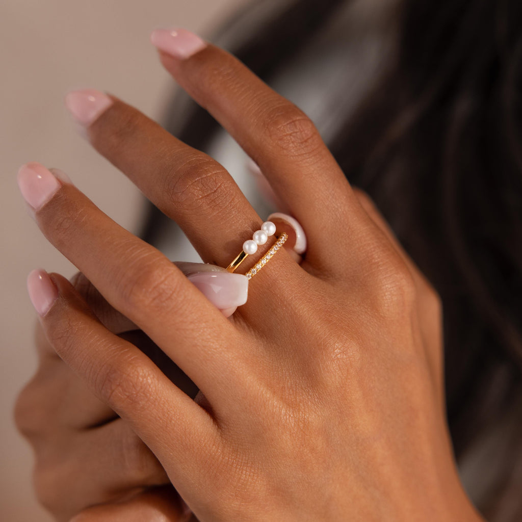 Close up of a woman's hand with light pink manicured nails, wearing a gold ring set featuring three small pearls and a slim pavé crystal band.