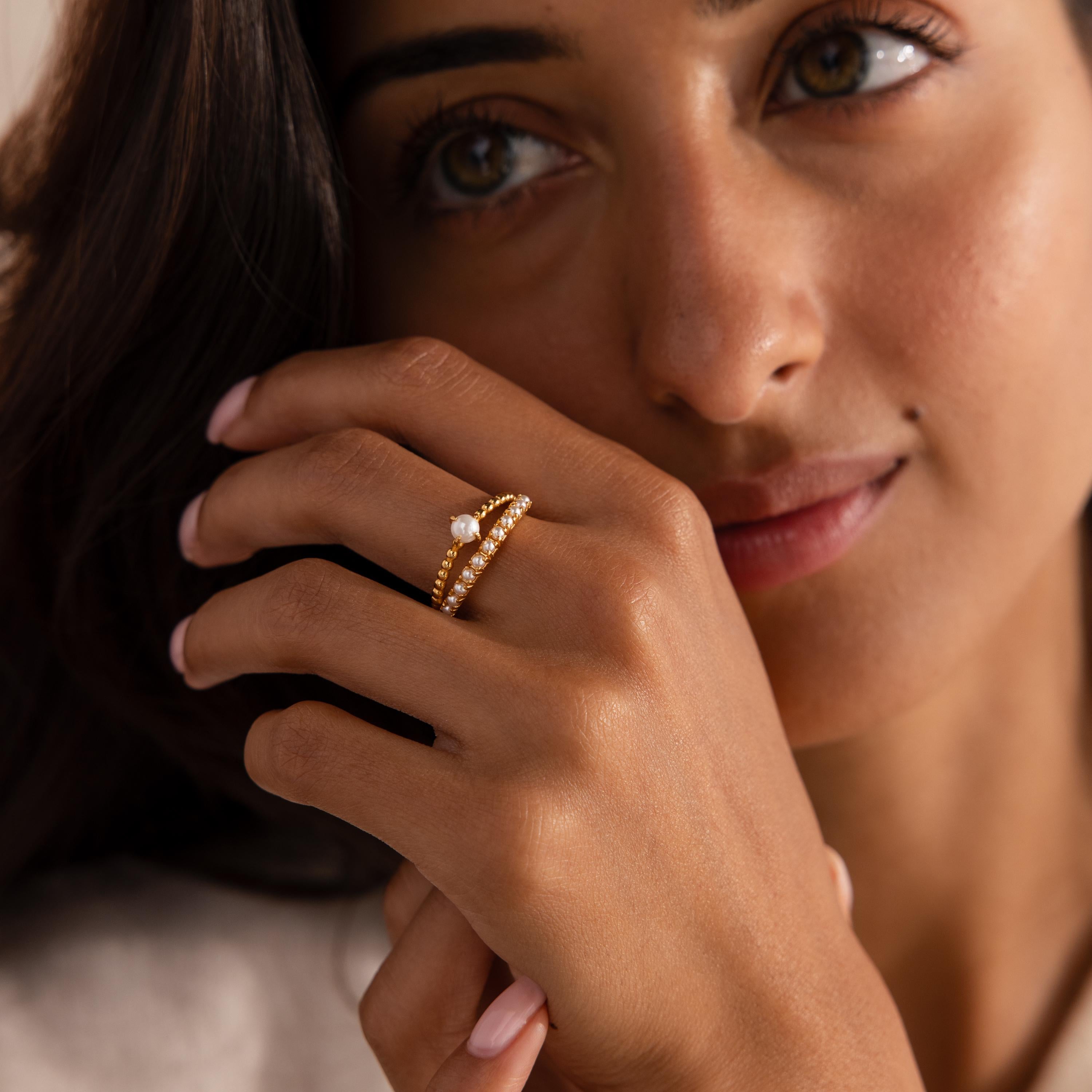 Woman wearing a gold ring set with beaded detailing, featuring a single central pearl on one band and a row of small pearls on the other.
