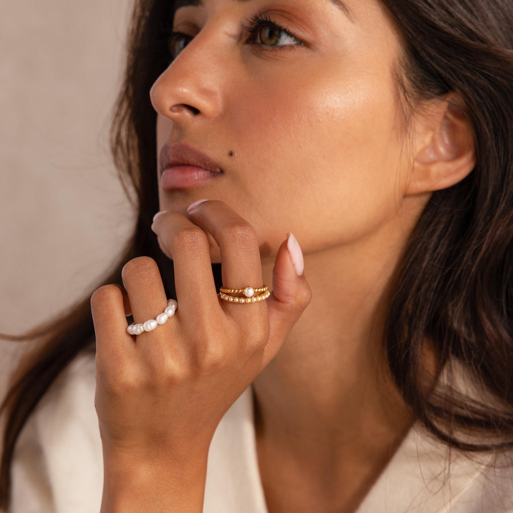 Woman wearing a gold beaded ring set with a central pearl and a row of small pearls, alongside a separate white beaded pearl ring, with her hand resting near her face.