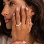 Close up of a woman’s hand against her cheek, wearing two rings: one strung with round white pearls and the other a gold beaded band featuring a central pearl with a row of tiny pearls along the lower band.