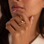 Woman wearing a gold beaded ring set, with one band featuring a single central pearl and the other adorned with small pearls, hand resting near her lips.