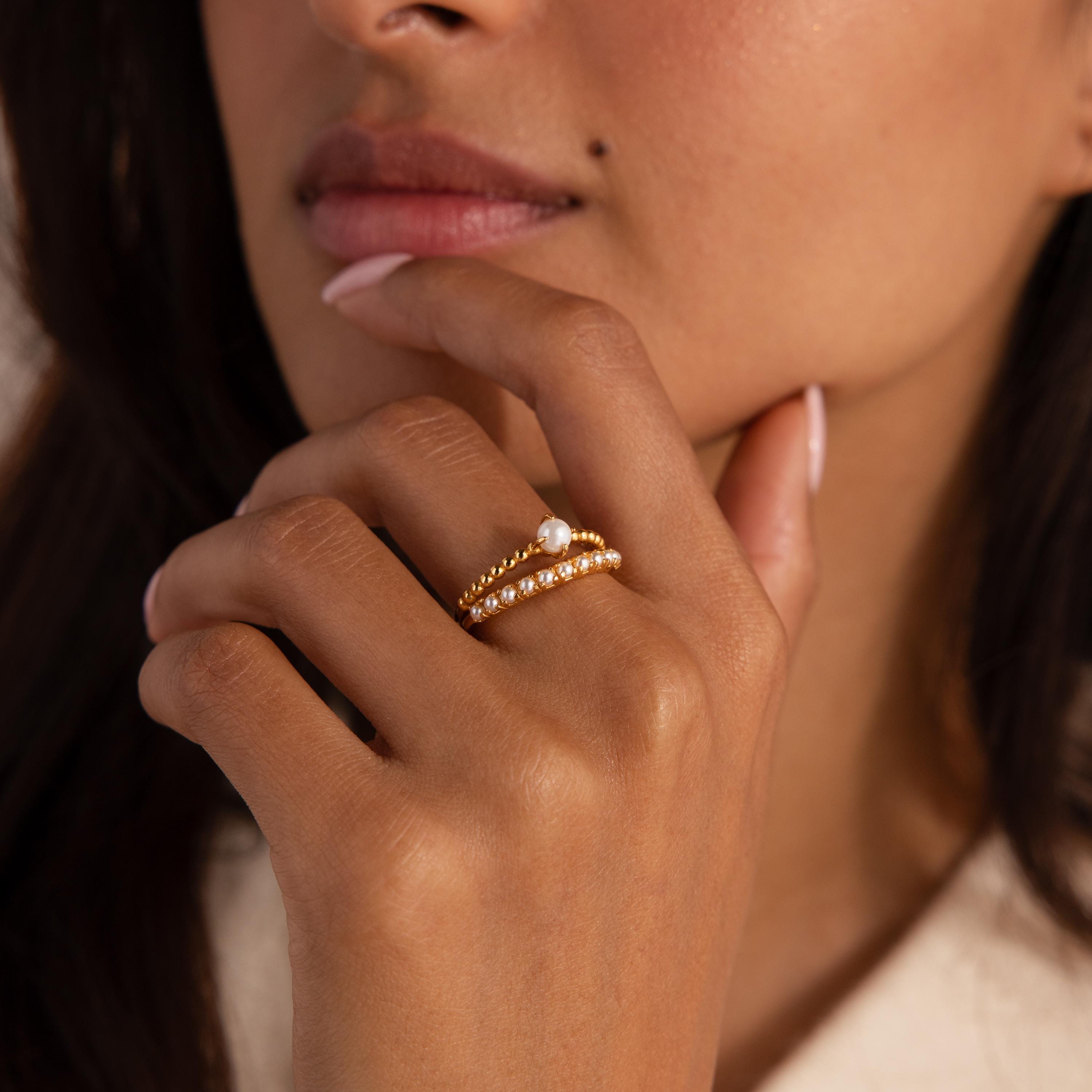 Woman wearing a gold beaded ring set, with one band featuring a single central pearl and the other adorned with small pearls, hand resting near her lips.
