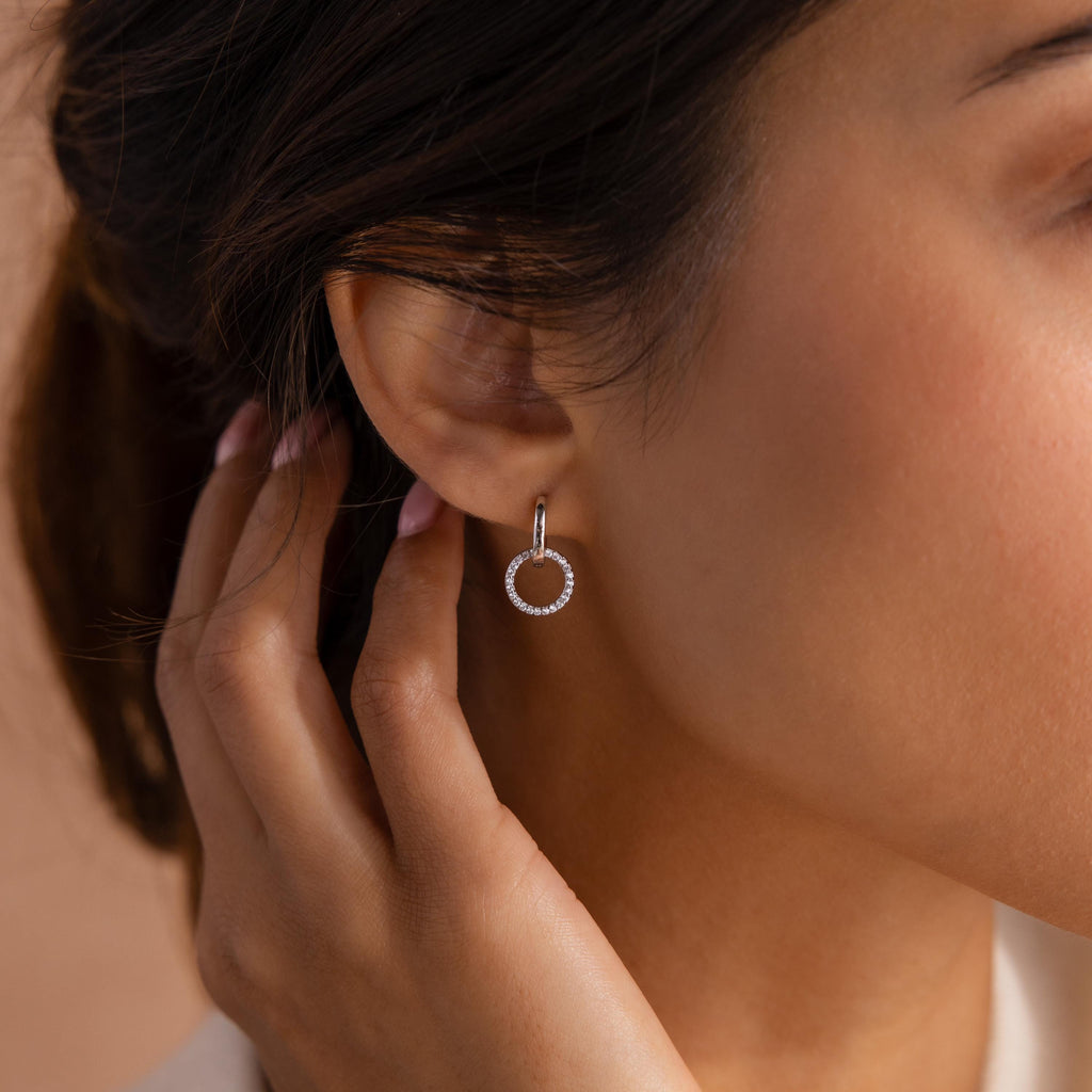 Woman wearing a silver hoop earring with a dangling open circle encrusted with small clear stones, touching her hair near her ear.