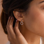 Woman wearing a silver hoop earring with a dangling open circle encrusted with small clear stones, touching her hair near her ear.