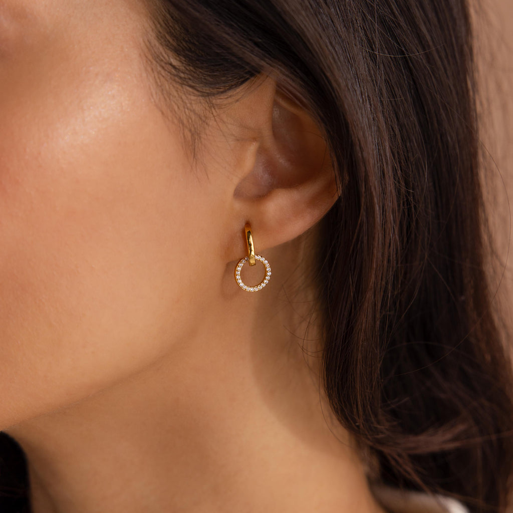 Gold hoop earring featuring a small dangling circle encrusted with clear stones, worn on the ear of a woman with long dark hair.