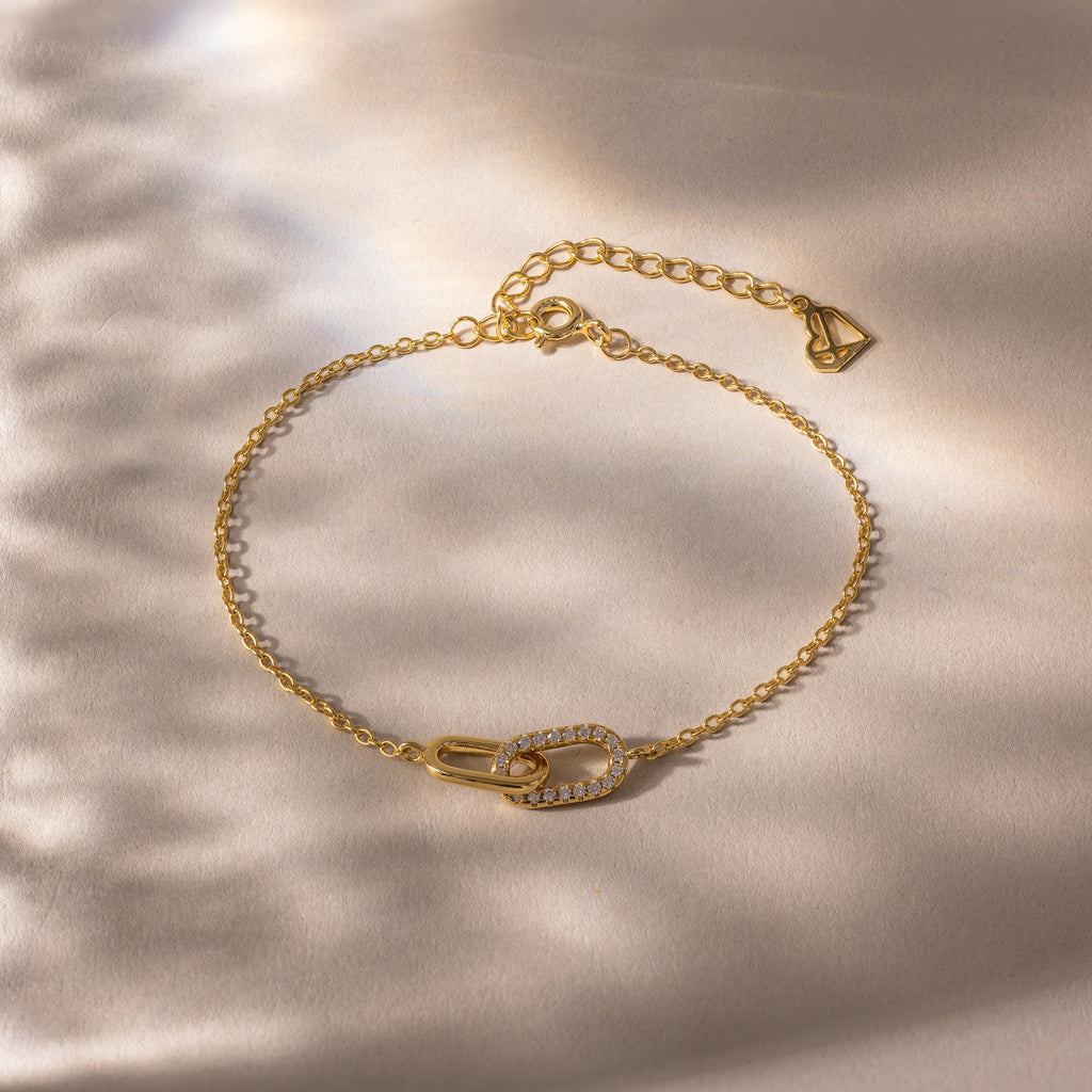 Delicate gold chain bracelet with two interlocking oval links at the center, one polished and one encrusted with small clear stones, displayed on a softly lit neutral background.