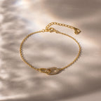 Delicate gold chain bracelet with two interlocking oval links at the center, one polished and one encrusted with small clear stones, displayed on a softly lit neutral background.