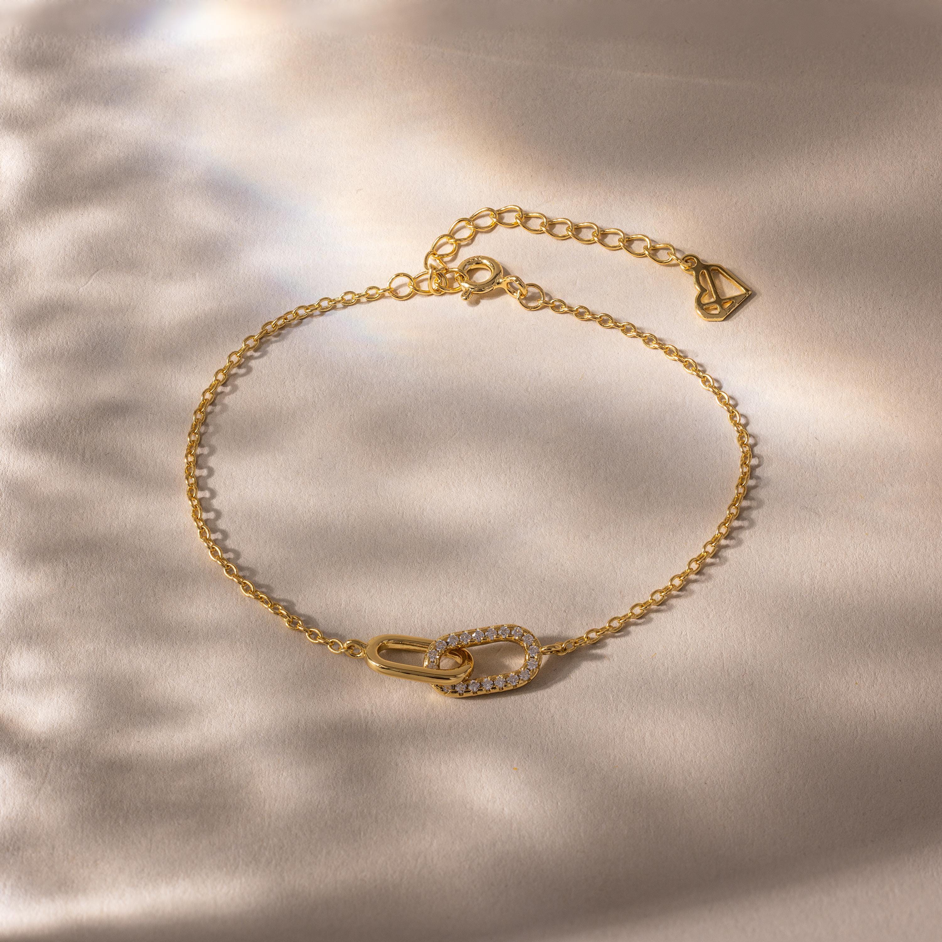 Delicate gold chain bracelet with two interlocking oval links at the center, one polished and one encrusted with small clear stones, displayed on a softly lit neutral background.