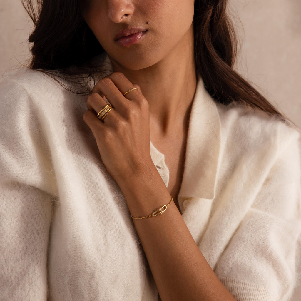 Woman wearing a cream knit top, gold stacking rings, and a delicate gold chain bracelet with two interlocking oval links, one polished and one adorned with small clear stones.