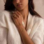 Woman wearing a cream knit top, gold stacking rings, and a delicate gold chain bracelet with two interlocking oval links, one polished and one adorned with small clear stones.