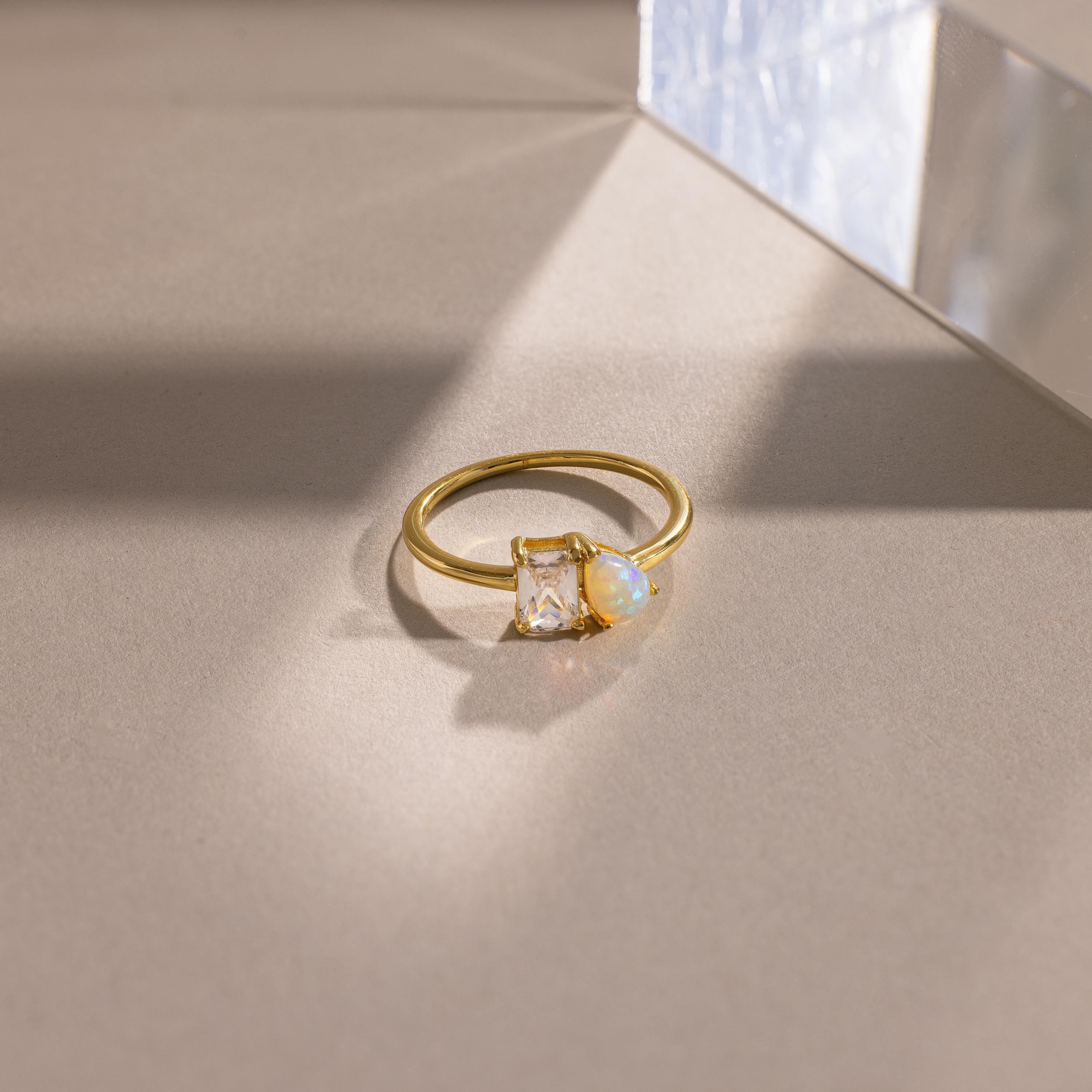 Gold ring featuring a rectangular clear gemstone paired with a pear-shaped opal, set side by side on a slim band, displayed on a smooth beige surface with geometric shadows.