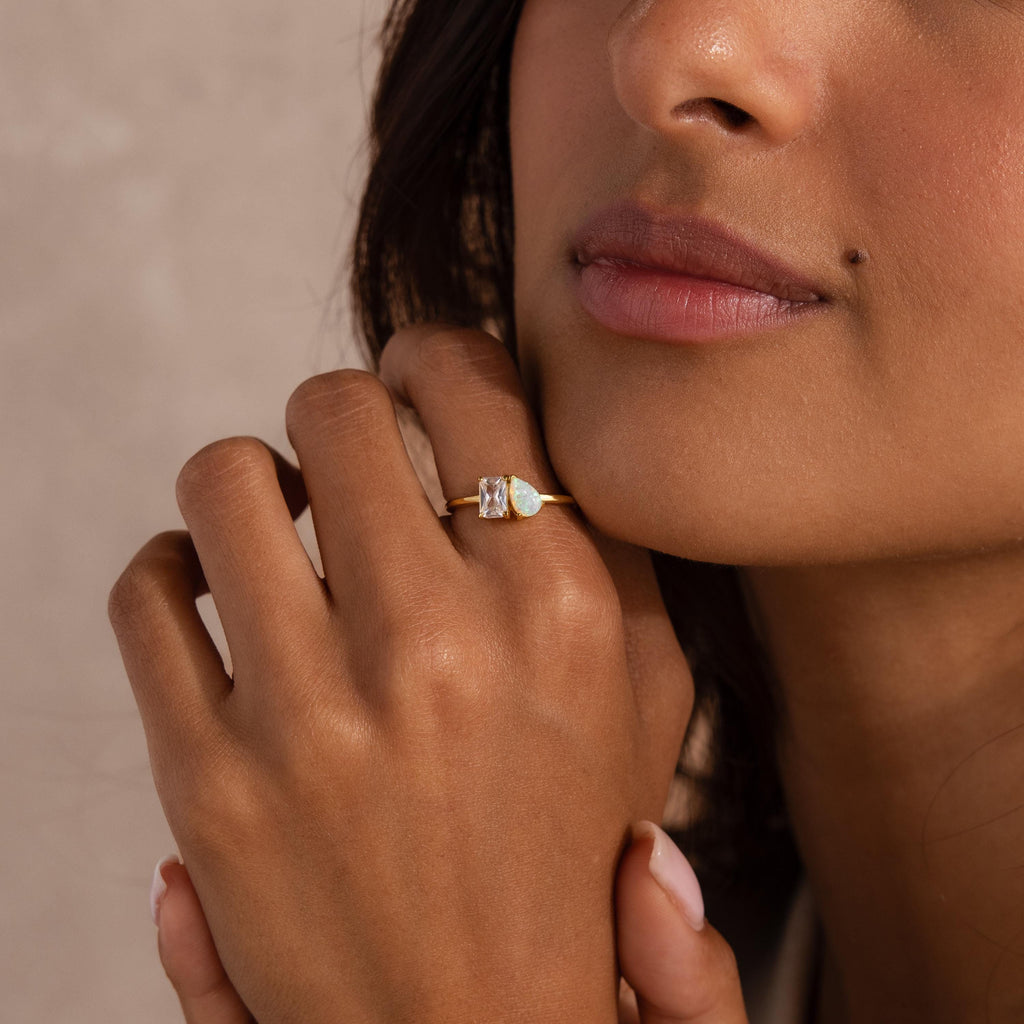 Close up of a woman’s hand near her chin, wearing a gold ring with a rectangular clear gemstone and a pear shaped opal set side by side.