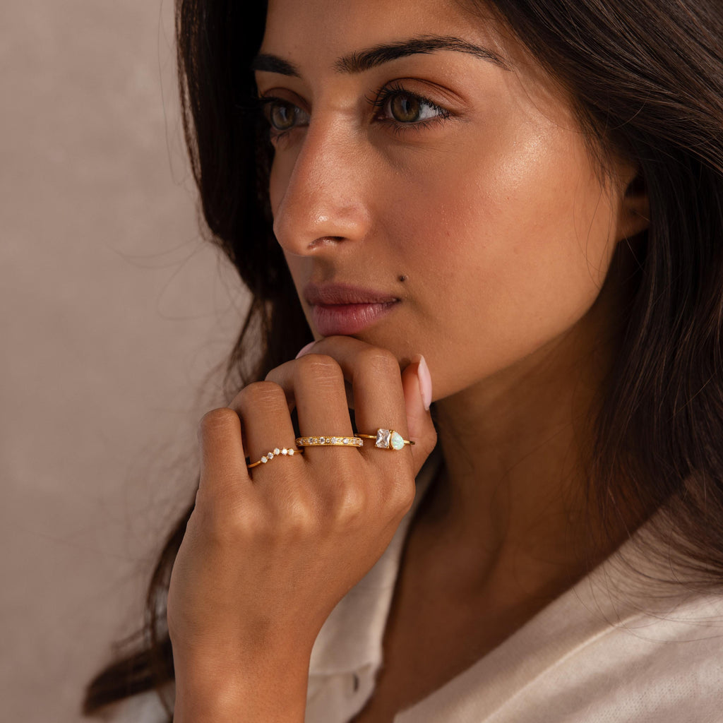 Woman with long dark hair wearing three gold rings, including one with a pear-shaped opal and rectangular clear gemstone, one with small pearls, and one with embedded clear stones.